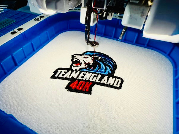 Embroidery machine with a 'Team England 40K' logo on a white fabric.